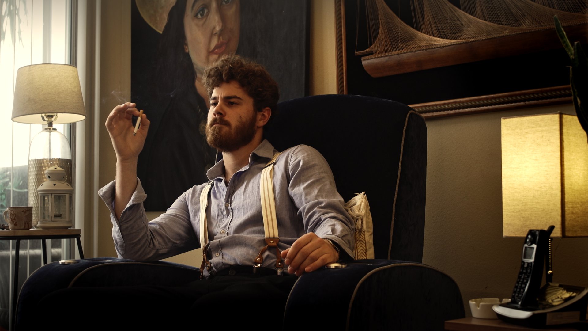 Still from God's Second Best Man: a man smoking in an armchair beside a lamp and a portrait
