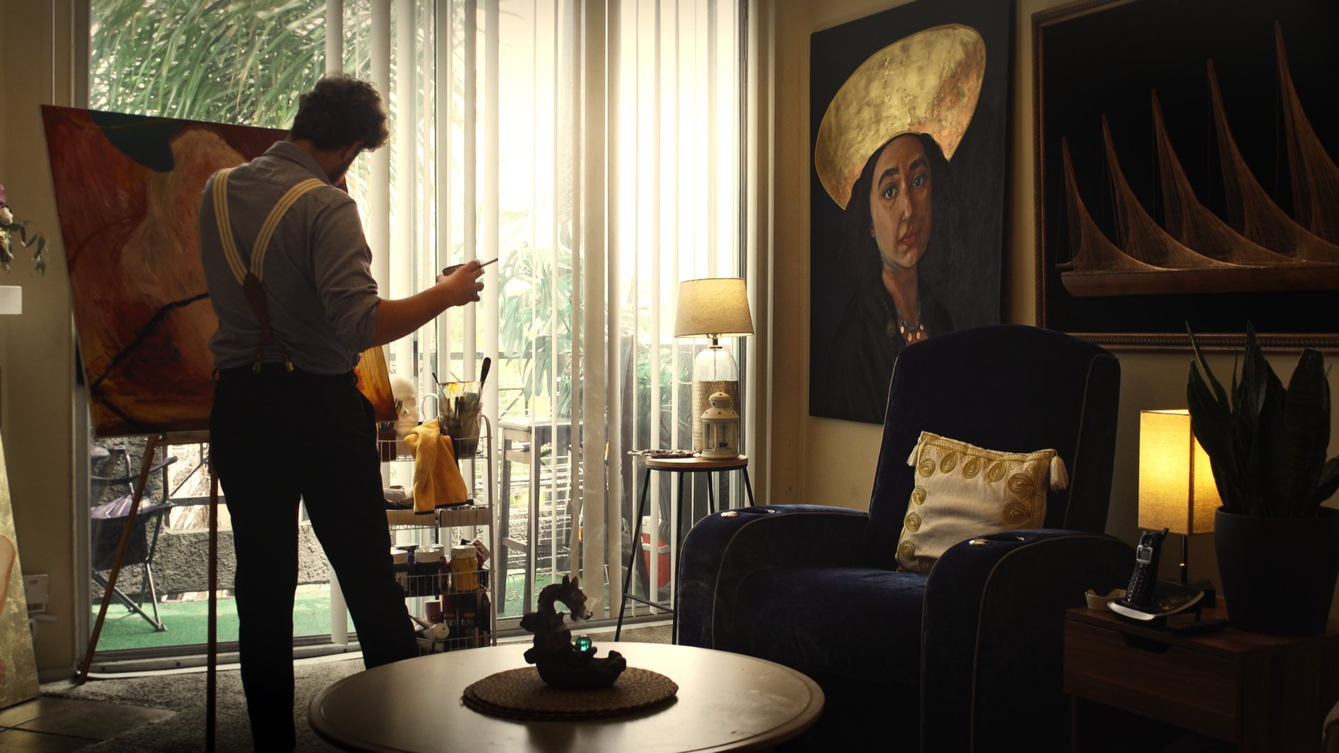 Still from God's Second Best Man: a man at a bright window surrounded by paintings