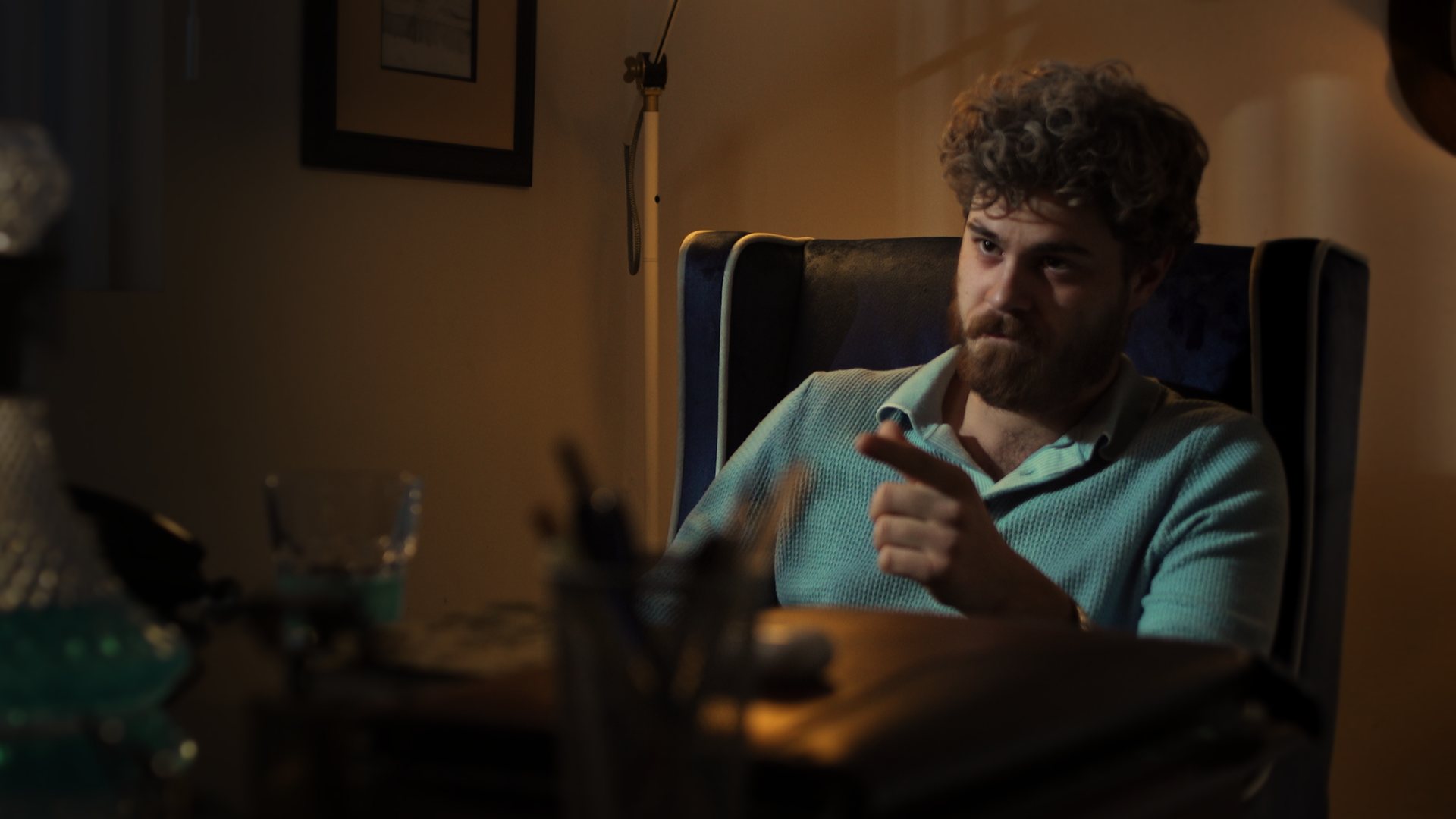 Still from God's Second Best Man: a man in a teal sweater talking across a desk