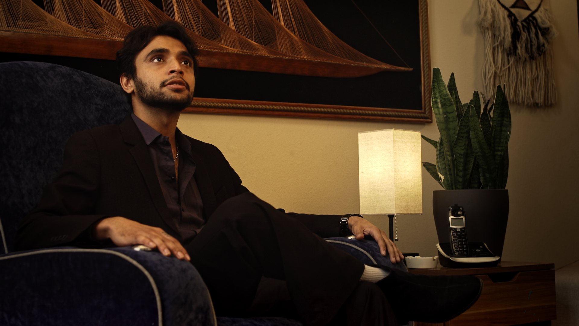 Still from God's Second Best Man: a man in a dark suit seated in an armchair, looking up