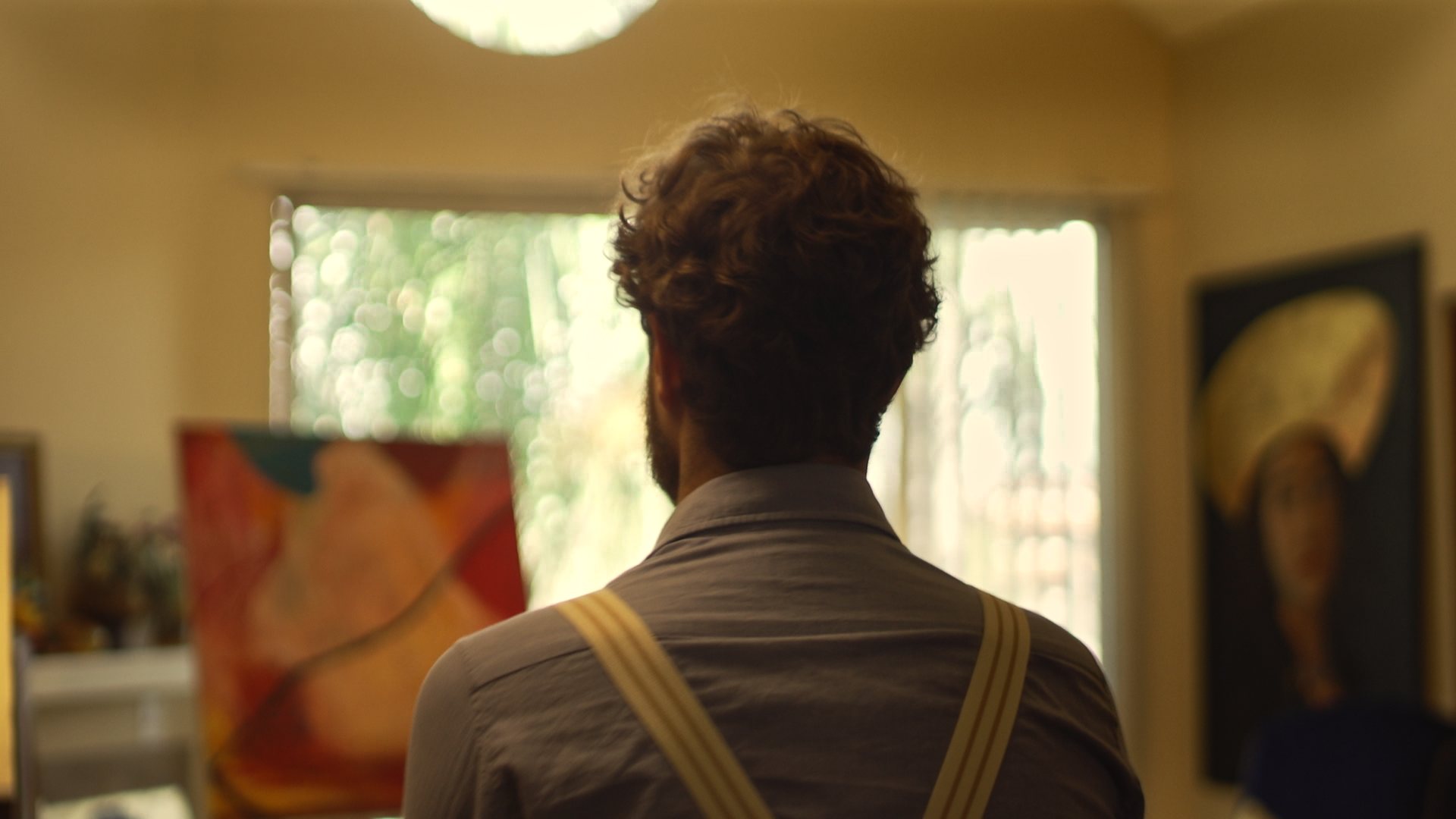 Still from God's Second Best Man: a man from behind, facing a bright window and paintings