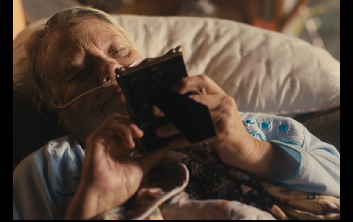 Still from What's Life: an elderly woman holding a camera in bed